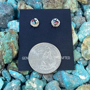 May include: A pair of small, round stud earrings with a silver-colored setting. Each earring features a central design with red, black, and white accents. The earrings are displayed on a black card next to a quarter coin. The card reads "GEN... CRAFTED".