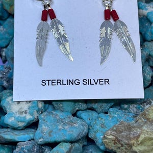 May include: Sterling silver earrings with a red stone top, red beads, and silver feather charms. The earrings are displayed on a white card with the words "STERLING SILVER". The background is a collection of turquoise stones.