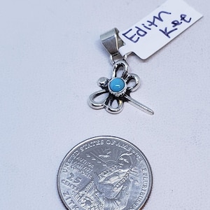 May include: A silver-toned pendant with a turquoise gemstone center, shaped like a stylized flower. The pendant is next to a U.S. quarter coin. A tag with the name "Edith Kee" is attached to the pendant.