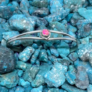 May include: A silver-toned bracelet with a pink gemstone center, set against a backdrop of turquoise stones. The bracelet features a double-band design and a round, pink gemstone. The turquoise stones vary in size and shade, creating a textured background.