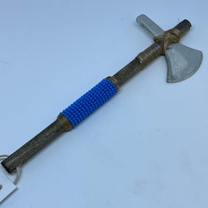 May include: A decorative tomahawk with a wooden handle and a silver-colored axe head. The handle features a section of bright blue beads and is wrapped with tan-colored twine.