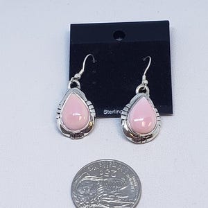 May include: A pair of sterling silver earrings with a teardrop-shaped, light pink stone. The earrings have a decorative silver border and a hook closure. The earrings are displayed on a black card with the word "Sterling" printed on it.