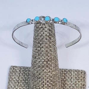 Puede incluir: Una pulsera de plata con cinco piedras turquesas. La pulsera se exhibe en un expositor de joyería texturizado de tono neutro. Las piedras turquesas están espaciadas uniformemente en la parte superior de la pulsera.