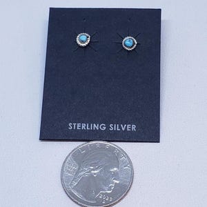 Puede incluir: Pendientes de botón de plata de ley con pequeñas piedras turquesas redondas engastadas en un borde plateado retorcido. Los pendientes se muestran en una tarjeta negra con la inscripción "STERLING SILVER". Una moneda de veinticinco centavos estadounidense está en primer plano.