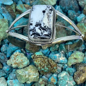 Può includere: Un bracciale a fascia in argento con una pietra rettangolare bianca venata di nero. La pietra è incastonata in un bordo a corda intrecciata e montata su una fascia a doppio gambo. Il bracciale ha una finitura metallica lucida.