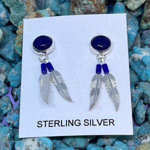 Può includere: Orecchini in argento sterling con una pietra blu scuro, perline blu pendenti e ciondoli a forma di piuma grigia. Gli orecchini sono esposti su una carta bianca con la scritta "STERLING SILVER". Lo sfondo è una collezione di pietre turchesi.