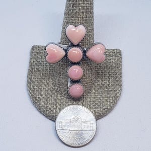 May include: A silver cross ring with pink heart and oval gemstones. The ring is shown on a ring holder next to a US nickel coin for size reference. The ring's design is reminiscent of a cross.