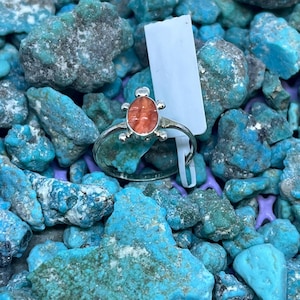 Op de afbeelding: Een zilveren ring met een schildpadvormige oranje steen, tegen een achtergrond van turkooizen stenen. De ring heeft een fijne band en een gedetailleerd schildpadontwerp, dat de kleur en vorm van de edelsteen benadrukt. De turkooizen stenen zorgen voor een levendig contrast.