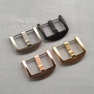 Könnte beinhalten: Vier Uhrenschnallen in verschiedenen metallischen Ausführungen: Silber, Schwarz, Roségold und Gold. Jede Schnalle hat einen rechteckigen Rahmen mit einer Mittelstange für das Uhrenarmband. Die Schnallen werden auf einer grauen Oberfläche präsentiert.