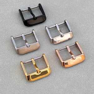 May include: An assortment of watch buckles in various metallic finishes. The buckles are in black, silver, rose gold, gold, and copper tones. Each buckle has a rectangular frame with a central bar for the watch strap.