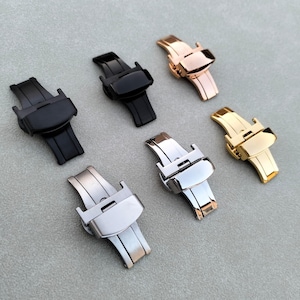 May include: An assortment of watch clasps in various metallic finishes, including black, silver, gold, and rose gold. The clasps are shown from a top-down perspective, highlighting their design and construction. The clasps are made of metal and have a butterfly clasp design.