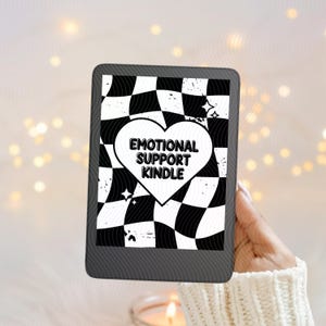 May include: A black e-reader shows a black and white chequered pattern with a white heart. The heart contains the text "EMOTIONAL SUPPORT KINDLE". The e-reader is held by a person wearing a white jumper.