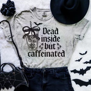 May include: A light grey t-shirt with the slogan "Dead inside but caffeinated" and a graphic of a drink with a straw. The shirt is styled with black denim shorts, a black hat, and a bat-shaped handbag.