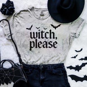 May include: A grey t-shirt with the words "Witch, Please" and bat graphics. The shirt is styled with black denim shorts, a black hat, and a bat-shaped handbag. A black scrunchie and bat cutouts complete the Halloween theme.