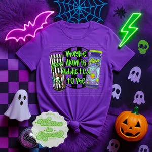 May include: Purple t-shirt with a graphic that says "Maybe Alani is addicted to me" with two cartoon beverage cans. The shirt is decorated with Halloween-themed elements like ghosts, a jack-o'-lantern, and a neon bat.
