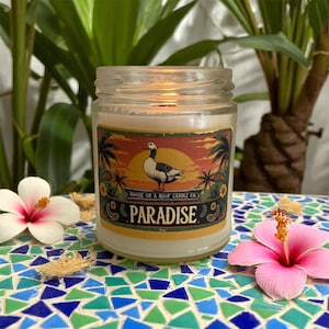 Puede incluir: Una vela encendida en un tarro de cristal con una etiqueta que dice "Paradise". La etiqueta presenta un ganso, palmeras y un diseño de puesta de sol. La vela está sobre una superficie de mosaico, con flores rosas y blancas en primer plano.