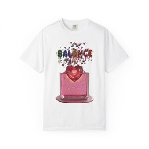 Op de afbeelding: Wit T-shirt met een grafisch ontwerp. Het ontwerp toont het woord "BALANCE" in kleurrijke, druipende letters boven een roze, hartvormig object in een gebarsten, roze vierkant. Er is ook een regenboog opgenomen.