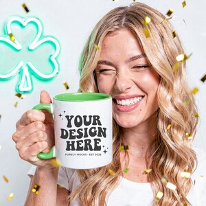 May include: White ceramic mug with a green handle and interior, featuring the text "YOUR DESIGN HERE". A neon green shamrock is in the background. Gold confetti is scattered around the mug. The mug is held by a person.