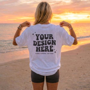 Op de afbeelding: Wit T-shirt met de tekst "YOUR DESIGN HERE" in het zwart, met sterren en de tekst "PURELY MOCKS EST 2025". Het shirt wordt gedragen op een strand bij zonsondergang. De persoon draagt een donkere korte broek.