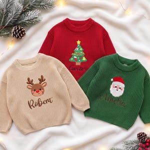 May include: Three long-sleeve knit sweaters in red, beige, and green. The red sweater features a Christmas tree and the name Carter. The beige sweater displays a reindeer and the name Robert. The green sweater shows Santa Claus and the name Amelia.