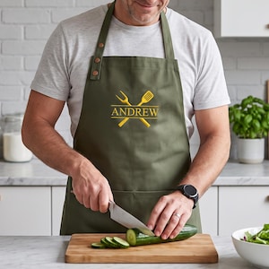 May include: An olive green apron featuring an embroidered yellow graphic of a crossed fork and spatula with the name ANDREW centered between them. The apron has two copper-colored metal buttons on the right shoulder strap and a front pocket.