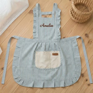 May include: A blue and white striped apron with a ruffled edge and a large front pocket. The apron has the name "Amelia" embroidered in brown thread. The apron has long ties and a neck strap. A small woven basket is in the background.