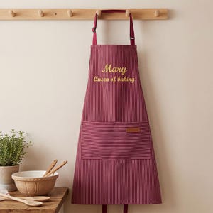 Può includere: Grembiule a righe bordeaux e bianche con il nome "Mary" e la frase "Queen of baking" ricamati in oro. Il grembiule ha una tasca frontale ed è appeso a un attaccapanni di legno.