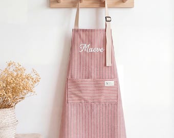 May include: A red and white striped apron with the name "Maeve" embroidered in white script. The apron has a large front pocket and is hanging on a wooden rack. The apron is made of a soft fabric.