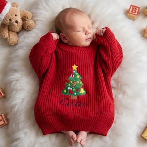May include: A red long-sleeved knit sweater featuring a green Christmas tree appliqué with a yellow star, ornaments, and the embroidered name Carter in black script. The sweater has a ribbed texture and a round neckline.