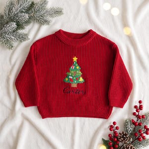 May include: A bright red long-sleeve knit sweater featuring a central embroidered Christmas tree design with a yellow star, colorful ornaments, and the name Carter stitched in dark thread below the tree trunk on the front.