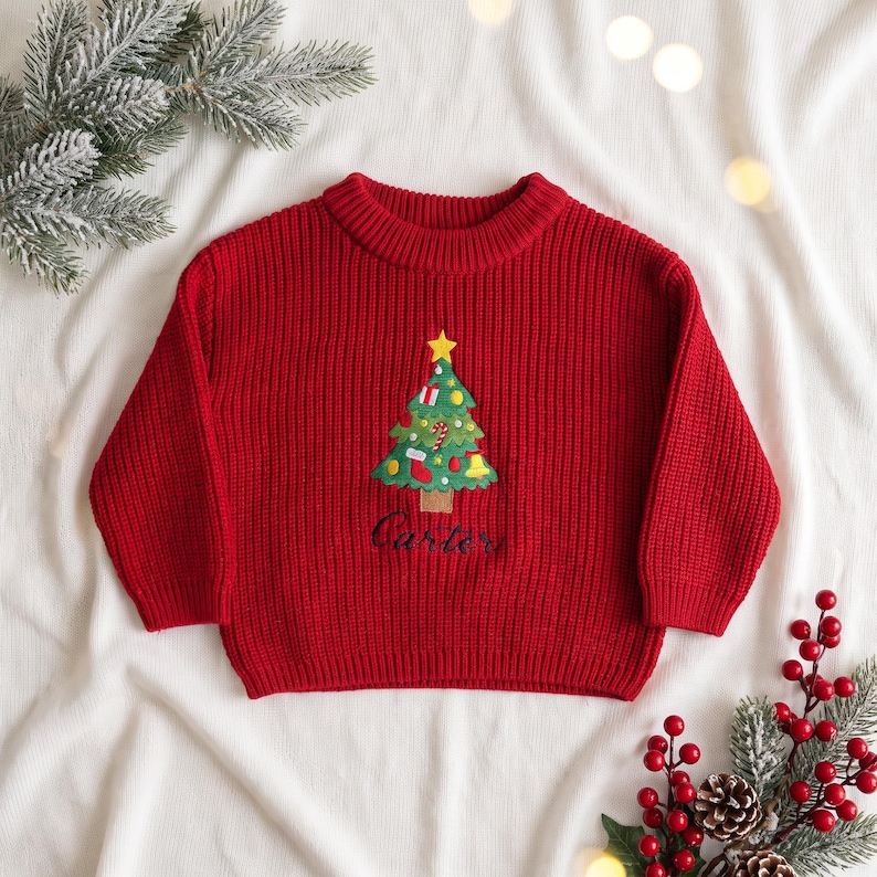 May include: A bright red long-sleeve knit sweater featuring a central embroidered Christmas tree design with a yellow star, colorful ornaments, and the name Carter stitched in dark thread below the tree trunk on the front.