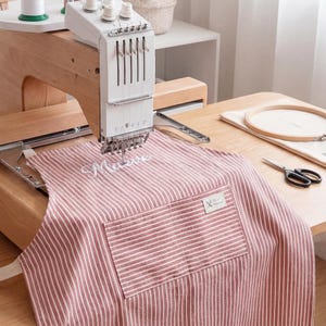 May include: An embroidery machine stitching the name "Maeve" onto a red and white striped apron. The apron has a pocket and a small label. A wooden embroidery hoop and scissors are on the wooden table.