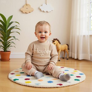 May include: A smiling baby wearing a tan sweater and pants set with the name "Hayden" embroidered on the front. The baby is sitting on a round rug with colorful polka dots. A toy horse and decorative clouds are in the background.
