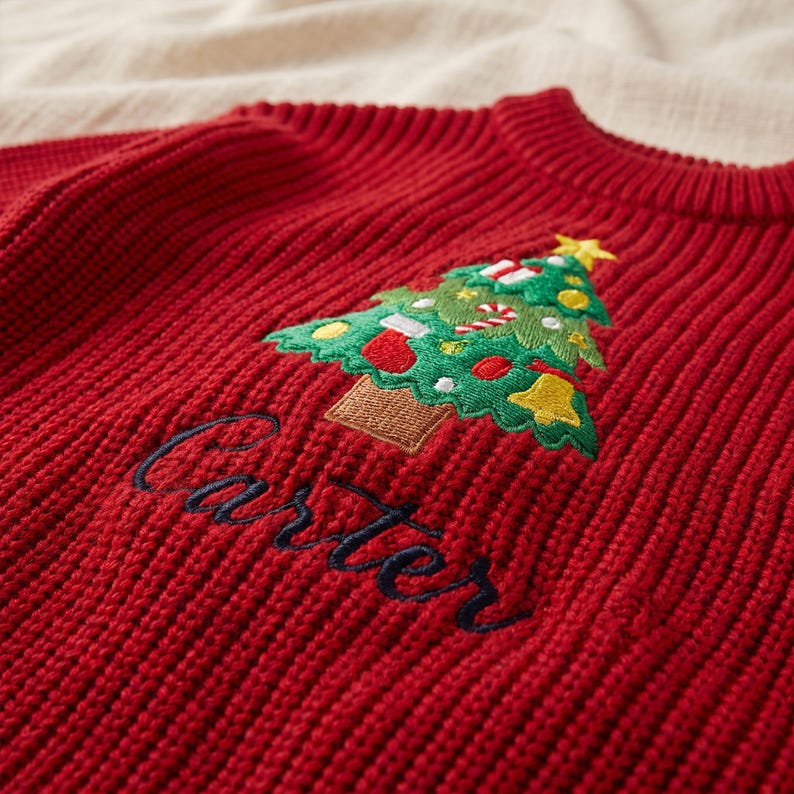 May include: A red ribbed knit sweater featuring an embroidered green Christmas tree decorated with colorful ornaments, a candy cane, and a yellow star. The name Carter is embroidered in dark blue cursive script below the tree.