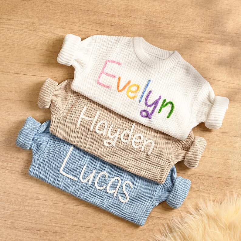 May include: Three personalized knit sweaters in white, tan, and light blue. The white sweater has the name "Evelyn" embroidered in rainbow colors. The tan sweater says "Hayden" and the blue sweater says "Lucas" in white.