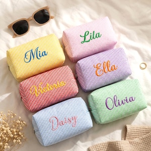 May include: A collection of colorful, striped cosmetic bags, each embroidered with a different name: Mia, Lila, Ella, Victoria, Olivia, and Daisy. The bags are in shades of yellow, pink, purple, red, green, and blue. Sunglasses are in the background.
