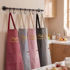 Könnte beinhalten: Fünf Schürzen mit vertikalen Streifen in Rot, Blau, Rosa, Schwarz und Grau hängen an einer schwarzen Metallstange. Jede Schürze hat eine Tasche und den Text "Mayah best pizza maker" in Gold.