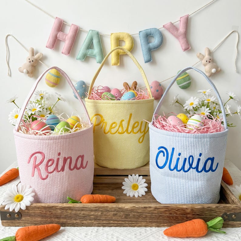 May include: Three Easter baskets in pink, yellow, and blue, each with a handle and a name embroidered on the front. The baskets are filled with colorful eggs and decorative grass. A banner that says "HAPPY" hangs in the background.
