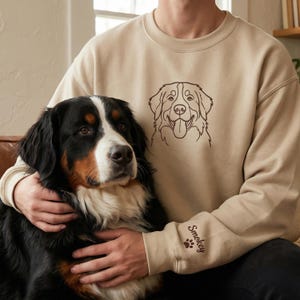 May include: A beige crewneck sweatshirt featuring a dark brown line art illustration of a dog on the chest. The left sleeve cuff displays the name Smokey in dark brown cursive script accompanied by a small dark brown paw print graphic.