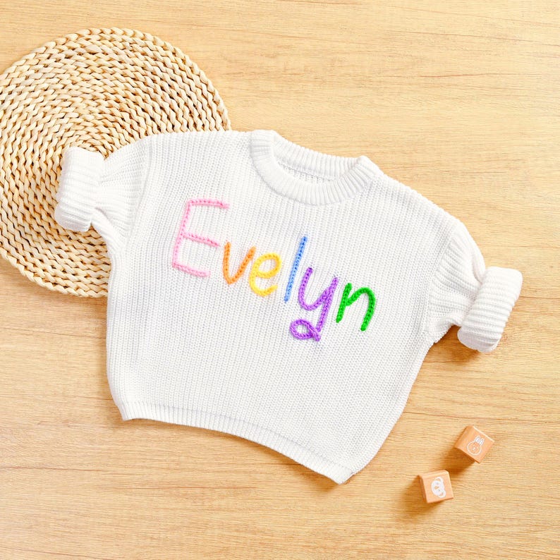 May include: White knit sweater with the name "Evelyn" embroidered in rainbow colors. The sweater has a crew neck and rolled-up sleeves. Two wooden blocks with letters are next to the sweater.