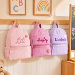 May include: Three striped backpacks hanging on a wooden rack. The left is light pink with the name Lili and a bow. The center is magenta with the name Hayley and a paw print. The right is lavender with the name Elizabeth and a daisy flower.