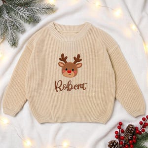 May include: A beige long-sleeve knit sweater featuring an embroidered brown reindeer with a red nose centered on the chest. Below the reindeer, the name Robert is embroidered in brown script lettering. The sweater has a ribbed crew neckline.