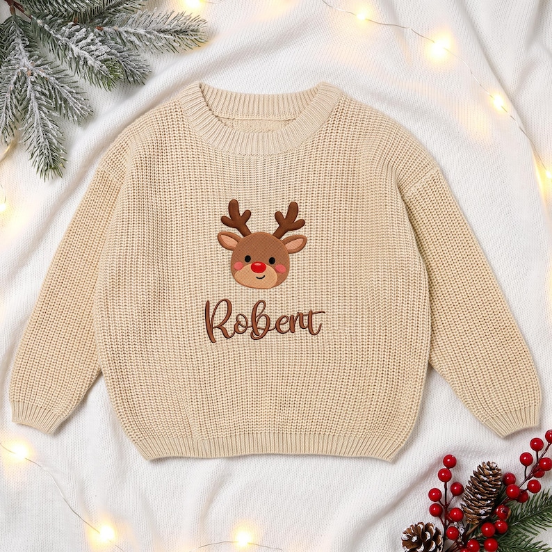 May include: A beige long-sleeve knit sweater featuring an embroidered brown reindeer with a red nose centered on the chest. Below the reindeer, the name Robert is embroidered in brown script lettering. The sweater has a ribbed crew neckline.