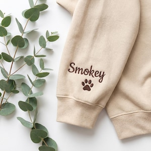 May include: A beige sweatshirt sleeve featuring dark brown embroidery on the cuff. The embroidery consists of the name Smokey in a cursive script font, positioned directly above a small, detailed dark brown paw print icon.