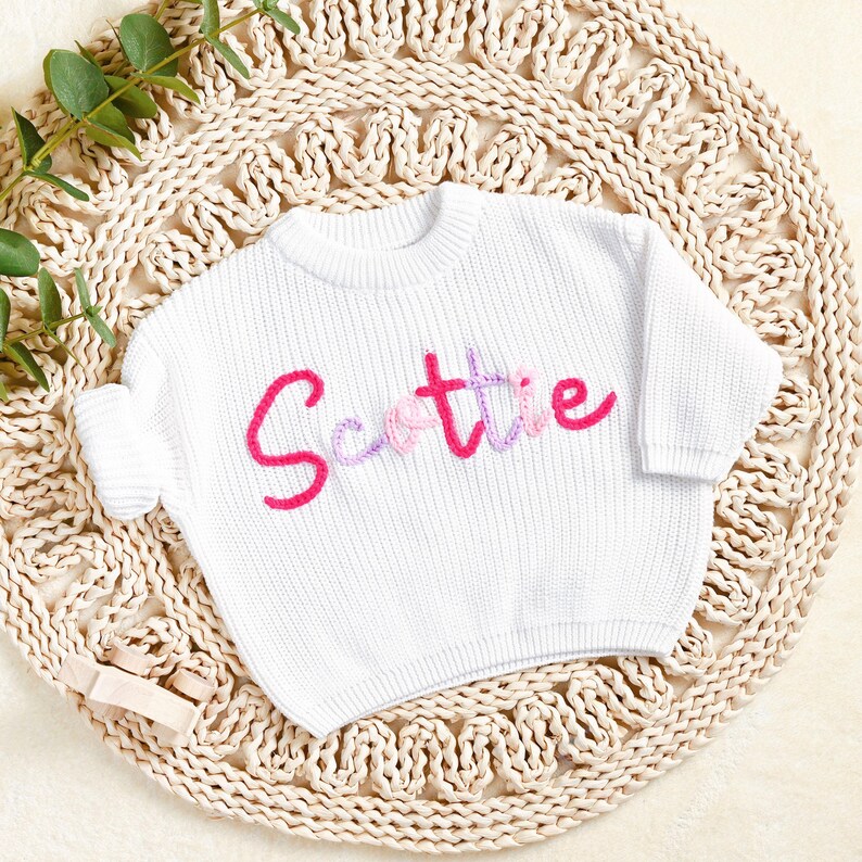 May include: White knit sweater with the name "Scottie" embroidered in pink and purple. The sweater is displayed on a woven, circular mat. A small wooden toy is visible in the lower left corner.