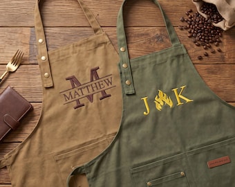 May include: Two canvas aprons, one tan and one olive green, featuring adjustable neck straps with metal rivets. The tan apron displays the embroidered name Matthew with a large letter M. The olive apron features the letters J and K with a flame icon.