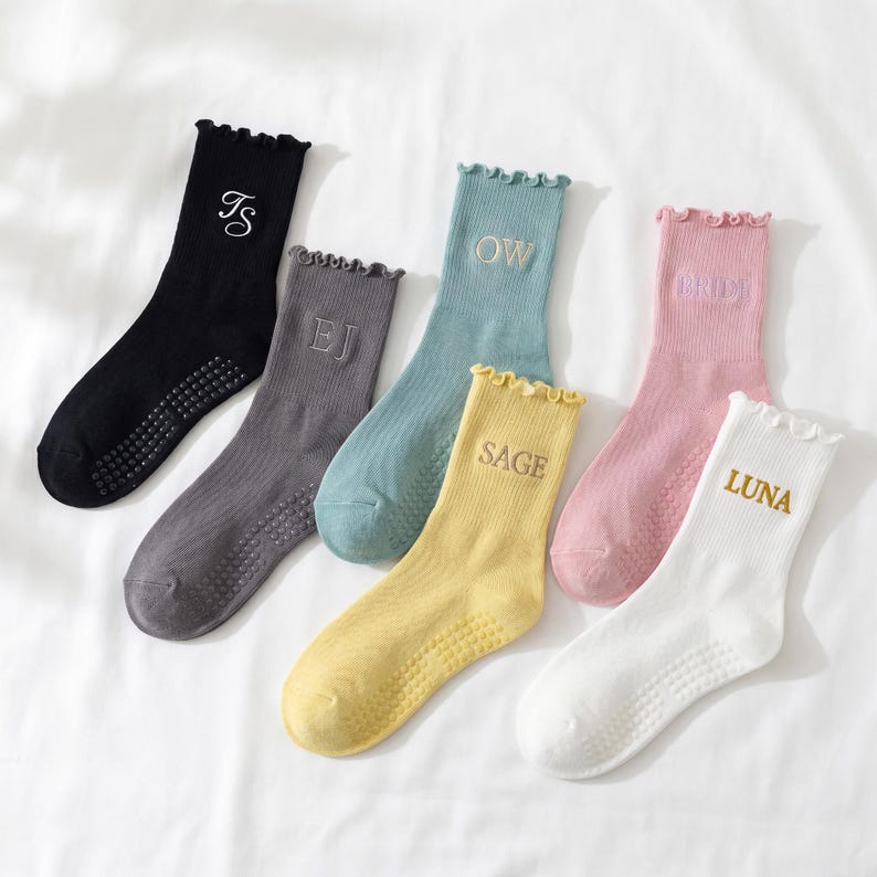 Puede incluir: Seis pares de calcetines en varios colores, incluyendo negro, gris, azul, amarillo, rosa y blanco. Cada calcet&iacute;n presenta un borde con volantes y iniciales o una palabra bordada. Los calcetines est&aacute;n dispuestos sobre una superficie blanca.