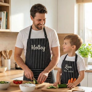 May include: A father and child wearing matching black and white striped aprons in a kitchen. The father's apron says "Holley" and the child's says "Liam Mini Artist". They are preparing food on wooden cutting boards.