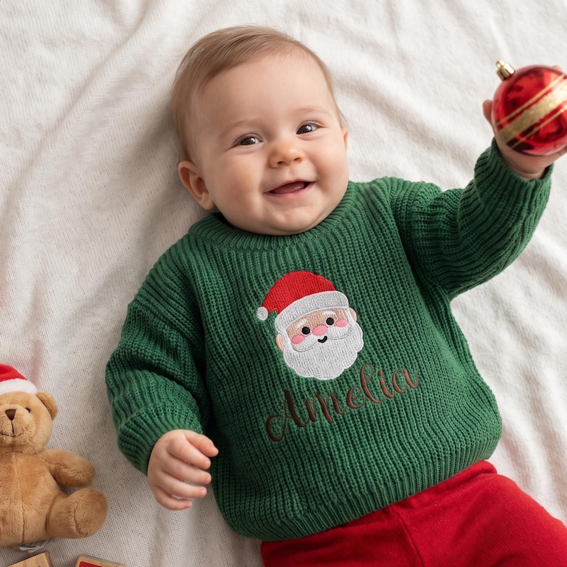 May include: A dark green long-sleeve knit sweater featuring an embroidered Santa Claus face and the name Amelia stitched in brown thread below the graphic. The sweater has a ribbed crew neckline, cuffs, and hem, worn with red pants.