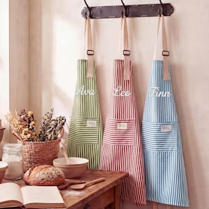 May include: Three striped aprons hang on a wall. The aprons are green, red, and blue, each with a white pocket and the names "Ava", "Leo", and "Finn" embroidered on them. The aprons have tan straps and are hanging from a dark metal rack.